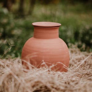 Op de afbeelding: Een terracotta pot met een deksel, die op een bed van stro staat. De pot is diep roodbruin van kleur en heeft een ronde vorm.