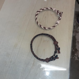 Puede incluir: Dos pulseras de cuero trenzado. Una es marrón y negra, la otra es blanca y marrón.
