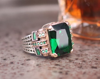 May include: A silver ring with a large, emerald green gemstone set in a gold-toned bezel. The ring band is braided and features smaller emerald green gemstones.