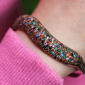 May include: A gold-toned bracelet with a curved design, encrusted with a multitude of colorful gemstones. The gemstones include shades of blue, green, orange, purple, and red. The bracelet is worn on a wrist, against a pink background.