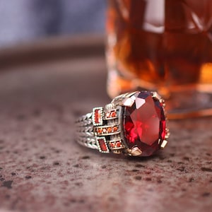 Könnte beinhalten: Ein silberner Ring mit einem großen, ovalen roten Edelstein. Der Ring ist mit kleineren roten Edelsteinen um den Hauptstein besetzt.