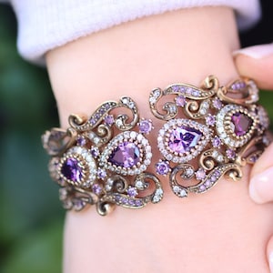 May include: A silver and gold bracelet with a delicate, ornate design. The bracelet is adorned with purple and clear gemstones.