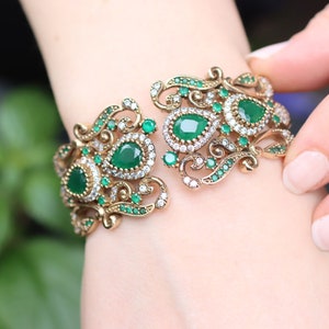 May include: A gold bracelet with intricate filigree design and green gemstones. The bracelet is open on one side and has a hinged clasp.