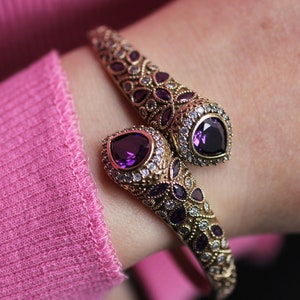 May include: A gold bangle bracelet with a snake design. The bracelet is adorned with purple and clear gemstones.