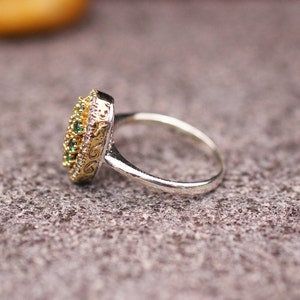 Pu&ograve; includere: Un anello d'argento con una montatura dorata e ornata. La montatura &egrave; incastonata con piccole pietre preziose verdi e bianche. L'anello &egrave; appoggiato su una superficie grigia strutturata.