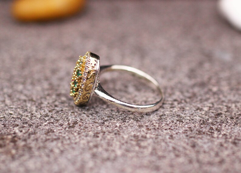 Pu&ograve; includere: Un anello d'argento con una montatura dorata e ornata. La montatura &egrave; incastonata con piccole pietre preziose verdi e bianche. L'anello &egrave; appoggiato su una superficie grigia strutturata.