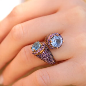 May include: A gold ring with two blue gemstones surrounded by purple stones.