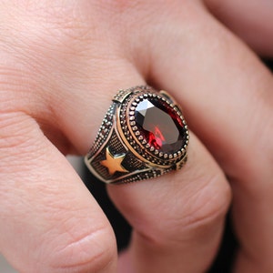 May include: A silver ring featuring a large, oval-shaped red gemstone set in a gold-toned bezel. The ring has a star design on the side.