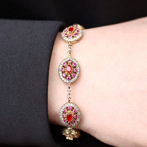 Może przedstawiać: Srebrna bransoletka z łańcuszkiem i trzema owalnymi zawieszkami. Każda zawieszka zdobiona jest czerwonym kamieniem szlachetnym otoczonym mniejszymi różowymi i białymi kamieniami szlachetnymi.