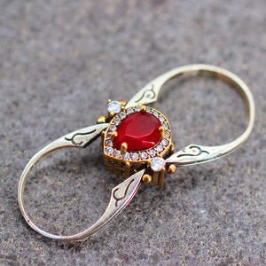 May include: A silver and gold ring with a red gemstone surrounded by smaller clear gemstones. The ring is designed to be worn on the thumb or finger.