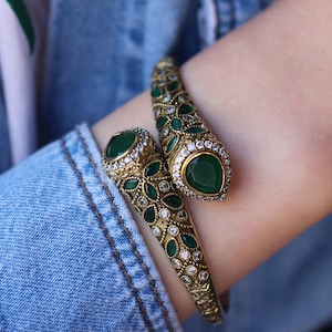 May include: A gold snake-shaped bangle bracelet with green and clear gemstones. The bracelet is on a person's wrist.