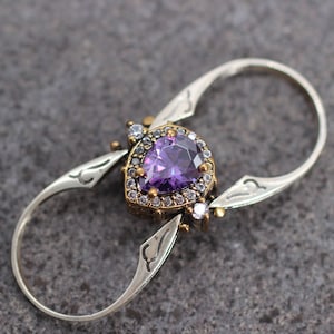 Op de afbeelding: Een zilveren ring met een paarse edelsteen omringd door kleinere heldere edelstenen. De ring heeft een uniek dubbelbandontwerp.