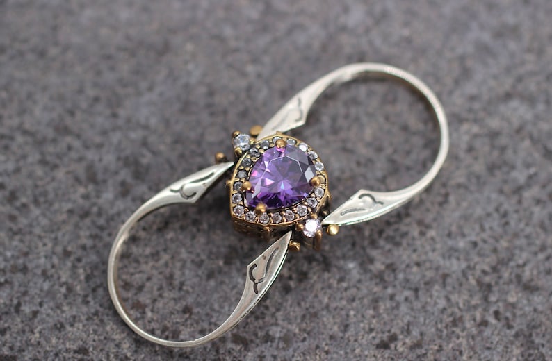 Op de afbeelding: Een zilveren ring met een paarse edelsteen omringd door kleinere heldere edelstenen. De ring heeft een uniek dubbelbandontwerp.