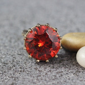 Peut inclure: Une bague avec une grande pierre pr&eacute;cieuse rouge facet&eacute;e sertie dans un m&eacute;tal dor&eacute;. La bague est pos&eacute;e sur une surface grise textur&eacute;e.