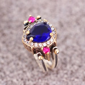 以下が含まれることがあります： 大きな青い涙型の宝石が、小さな白い宝石で囲まれた銀の指輪。指輪の側面には小さなピンクの宝石が2つある。