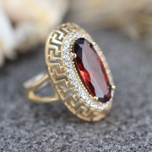 May include: A gold ring with a large, oval-shaped red gemstone surrounded by smaller white gemstones. The ring has a decorative, patterned band.