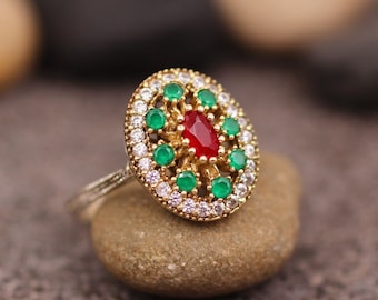 May include: A gold ring featuring a red and green gemstone setting. The ring has a round design with a central red stone surrounded by smaller green stones.