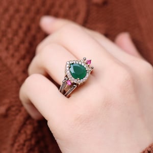 Könnte beinhalten: Ein silberner Ring mit einem birnenförmigen grünen Edelstein, der von kleineren klaren Steinen umgeben ist. Der Ring hat auch drei kleine rosa Steine auf der Ringschiene.