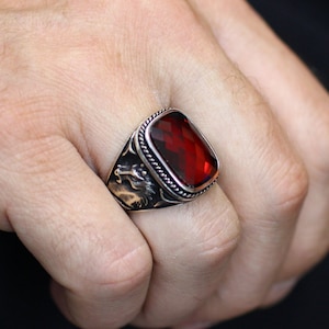 Puede incluir: Un anillo de plata con una gran piedra rectangular roja. El anillo tiene un diseño detallado en la banda, con una cabeza de animal estilizada.