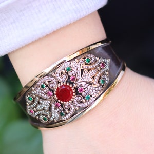 May include: A silver and gold bangle bracelet with an intricate floral design. The design features a large red gemstone surrounded by smaller green and pink gemstones.