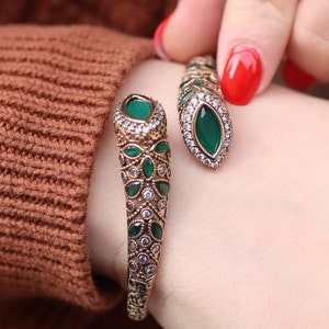 May include: A gold-toned bracelet with green stones and clear rhinestones. The bracelet is designed in a snake shape and is open on one side.