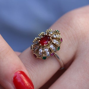 Peut inclure: Une bague en or avec une grande pierre rouge entourée de petites pierres blanches et des accents verts et rouges.