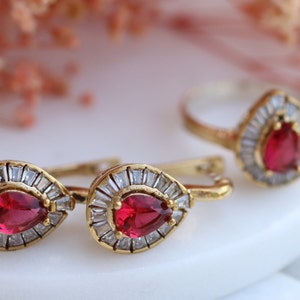 May include: A set of gold-tone jewelry featuring a teardrop-shaped red gemstone surrounded by clear cubic zirconia stones. The set includes a ring and a pair of earrings.