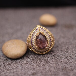 Puede incluir: Un anillo de oro con una gran piedra preciosa de color p&uacute;rpura en forma de l&aacute;grima rodeada de piedras m&aacute;s peque&ntilde;as de color claro y p&uacute;rpura.