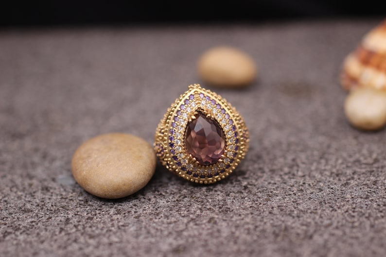 Puede incluir: Un anillo de oro con una gran piedra preciosa de color p&uacute;rpura en forma de l&aacute;grima rodeada de piedras m&aacute;s peque&ntilde;as de color claro y p&uacute;rpura.