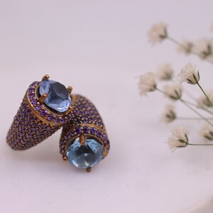 May include: Two gold rings with blue gemstones and purple accents. The rings are set on a white surface with white flowers in the background.