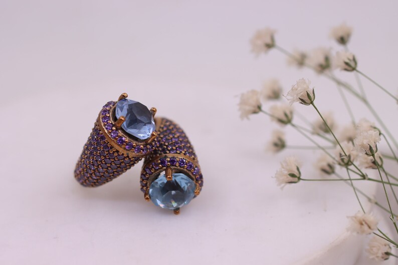 May include: Two gold rings with blue gemstones and purple accents. The rings are set on a white surface with white flowers in the background.