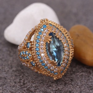 Puede incluir: Un anillo de oro con una gran piedra preciosa azul rodeada de piedras azules y blancas más pequeñas.