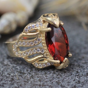 Op de afbeelding: Een gouden ring met een grote, rode, ovale edelsteen omringd door kleinere, heldere edelstenen.