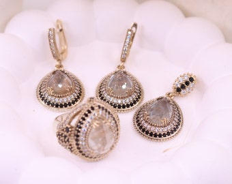 May include: A gold-tone jewelry set with a teardrop-shaped ring, earrings, and a pendant. Each piece features a clear stone surrounded by a border of small white and black stones.