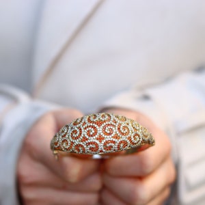 K&ouml;nnte beinhalten: Ein gold- und silberfarbenes Armband mit einem wirbelnden Muster aus orangefarbenen und wei&szlig;en Steinen.