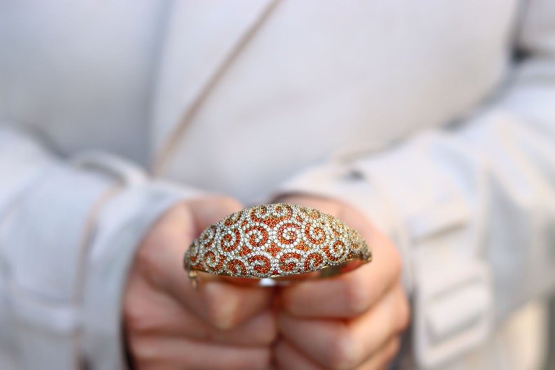 K&ouml;nnte beinhalten: Ein gold- und silberfarbenes Armband mit einem wirbelnden Muster aus orangefarbenen und wei&szlig;en Steinen.