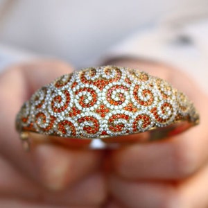 K&ouml;nnte beinhalten: Ein goldenes Armreif-Armband mit einem Wirbelmuster aus wei&szlig;en und orangefarbenen Edelsteinen.