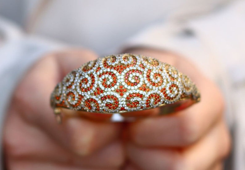 K&ouml;nnte beinhalten: Ein goldenes Armreif-Armband mit einem Wirbelmuster aus wei&szlig;en und orangefarbenen Edelsteinen.