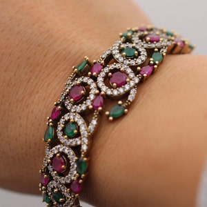 May include: A gold bracelet with an intricate design featuring pink and green gemstones. The bracelet is made of metal and has a delicate, feminine look.