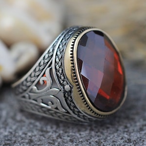 Può includere: Un anello d'argento con un designo ornato e una grande pietra rossa sfaccettata.