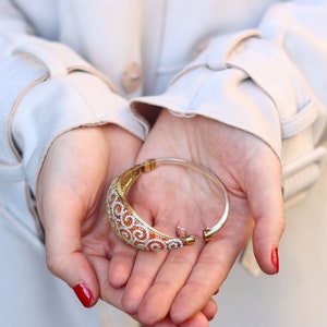 K&ouml;nnte beinhalten: Ein goldfarbenes und wei&szlig;es Armband mit einem Spiralmuster. Das Armband ist offen und liegt in den Handfl&auml;chen von zwei H&auml;nden.