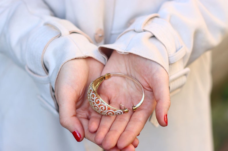 K&ouml;nnte beinhalten: Ein goldfarbenes und wei&szlig;es Armband mit einem Spiralmuster. Das Armband ist offen und liegt in den Handfl&auml;chen von zwei H&auml;nden.