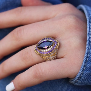 May include: A gold ring with a large, faceted, purple gemstone surrounded by smaller purple and white gemstones.