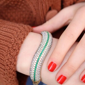 May include: A gold bangle bracelet with a curved design. The bracelet is set with small, clear stones and a row of green stones.