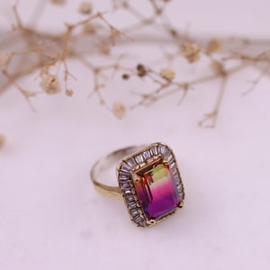 Puede incluir: Un anillo de plata y oro con una gran piedra rectangular con un degradado de colores de verde a rosa. La piedra preciosa est&aacute; rodeada de piedras m&aacute;s peque&ntilde;as, transparentes y de corte baguette. El anillo est&aacute; sobre una superficie blanca.
