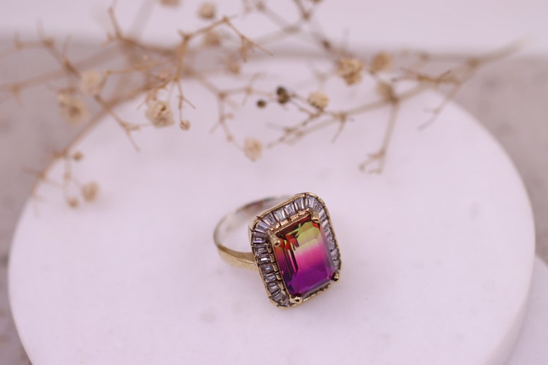 Puede incluir: Un anillo de plata y oro con una gran piedra rectangular con un degradado de colores de verde a rosa. La piedra preciosa est&aacute; rodeada de piedras m&aacute;s peque&ntilde;as, transparentes y de corte baguette. El anillo est&aacute; sobre una superficie blanca.
