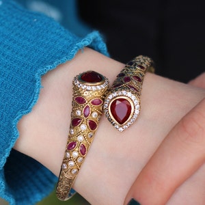 Peut inclure: Un bracelet serpent en or avec des pierres précieuses rouges et blanches. Le bracelet est au poignet d'une personne.