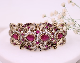 Pode incluir: Pulseira dourada ornamentada com pedras preciosas vermelhas em forma de gota e pedras redondas vermelhas e claras menores. A pulseira tem um design vazado com detalhes em rolagem, provavelmente uma joia.