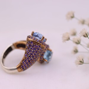 May include: A silver ring with a gold-toned setting featuring two blue gemstones surrounded by smaller purple gemstones.