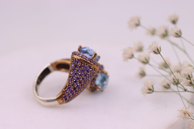 May include: A silver ring with a gold-toned setting featuring two blue gemstones surrounded by smaller purple gemstones.